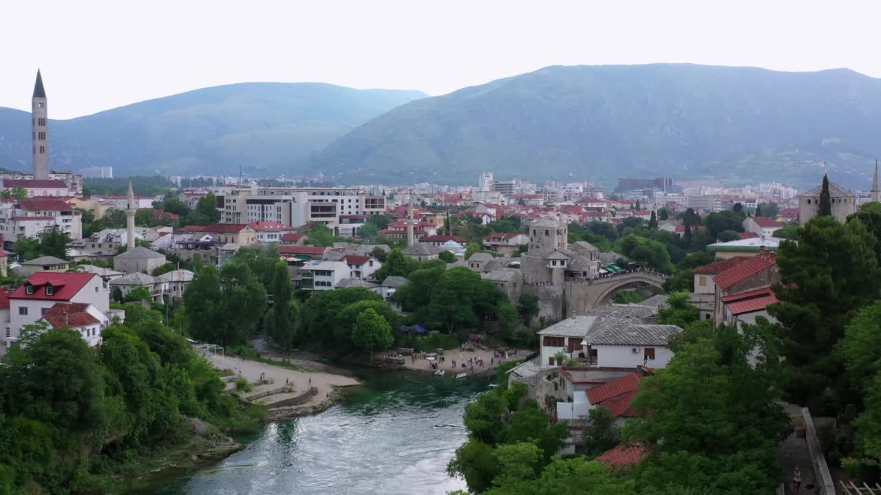 航拍:游客在河上探索古老的拱桥，无人机向后飞过绿树-莫斯塔尔，波斯尼亚和黑塞哥维那视频素材