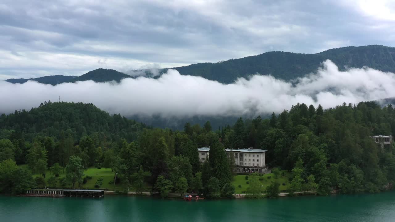 鸟瞰图:山上森林上空的云朵，无人机在绿色树木中的建筑上方飞行-流血，斯洛文尼亚视频下载