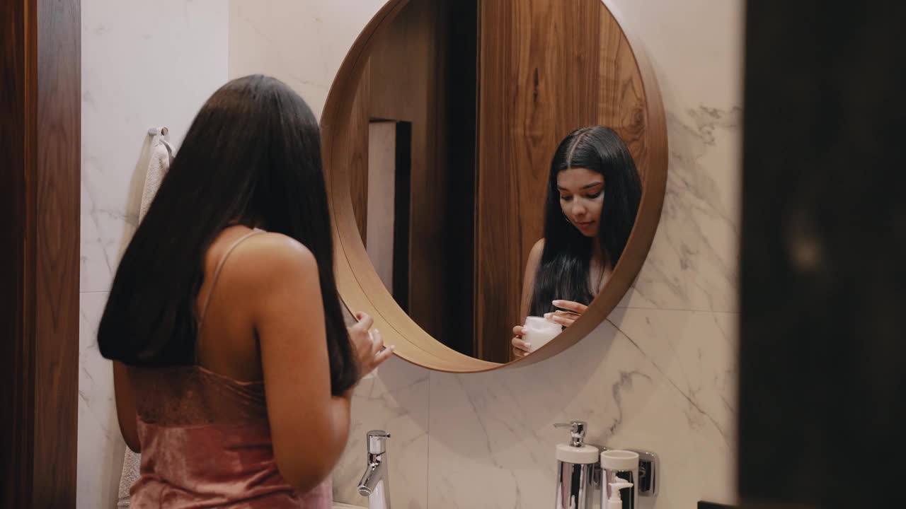 一个皮肤黝黑的年轻女子在镜子前给她的脸上涂奶油。视频素材