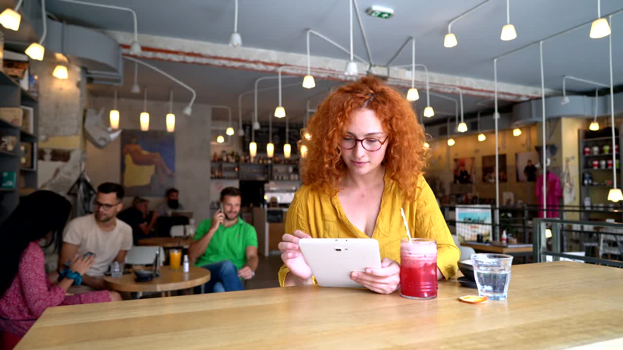 开朗的红发年轻女子，在现代舒适的自助餐厅通过电子平板电脑学习视频素材