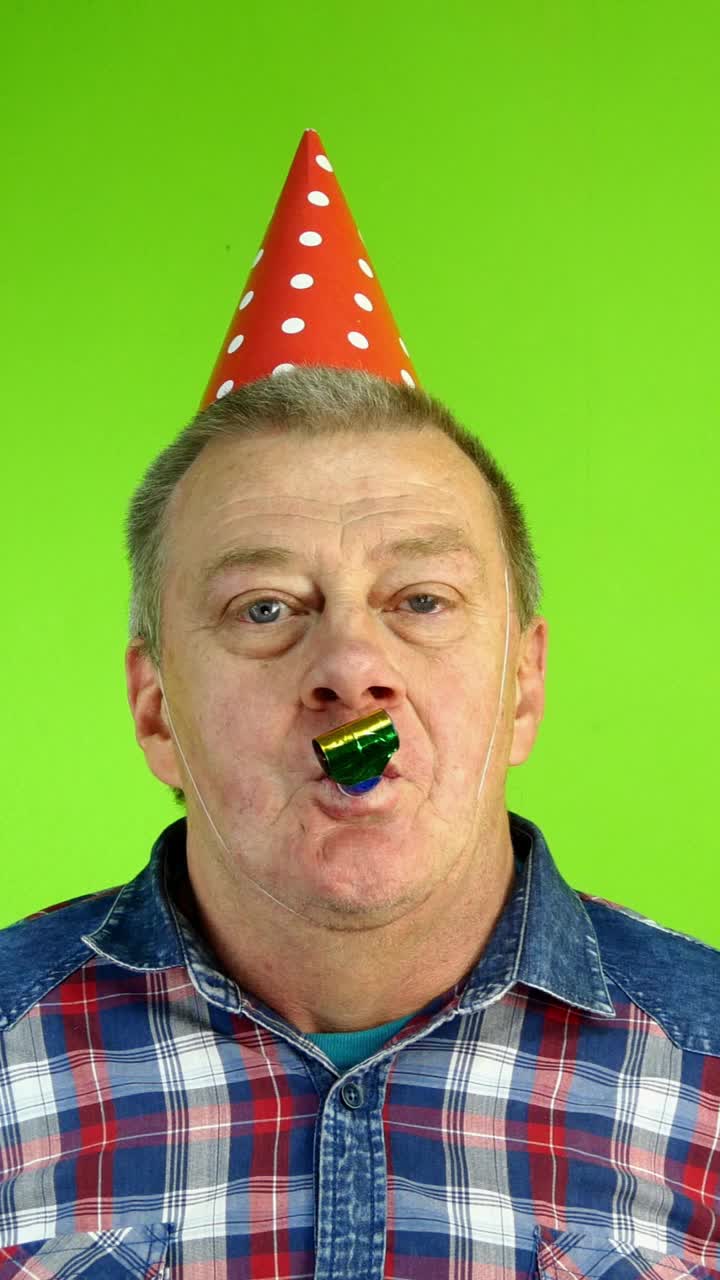 一名白人成年老人戴着生日帽，在眼睛上涂上五颜六色的甜甜圈，然后对着节日烟斗吹气。视频下载