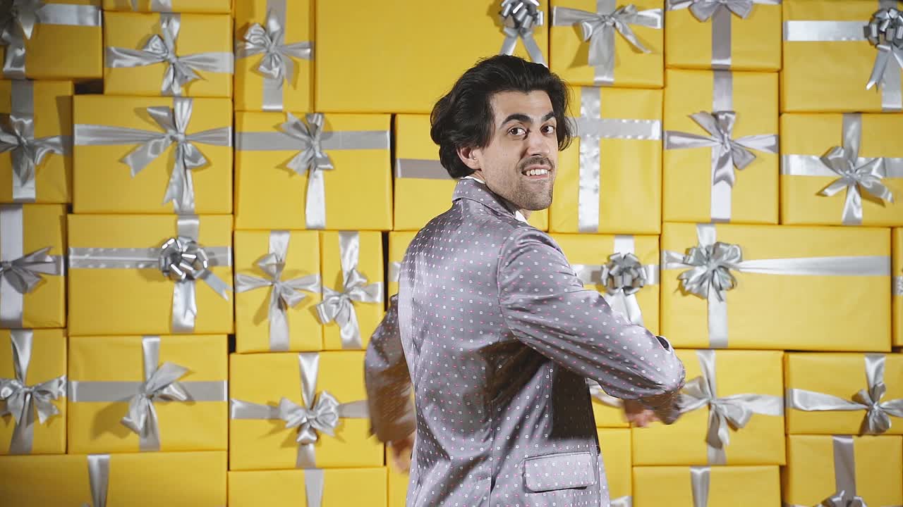 一个穿着优雅的灰色西装的开朗的年轻人在一间满是礼品盒的房间里跳舞，在礼物的背景下对着镜头摆姿势。圣诞节视频素材