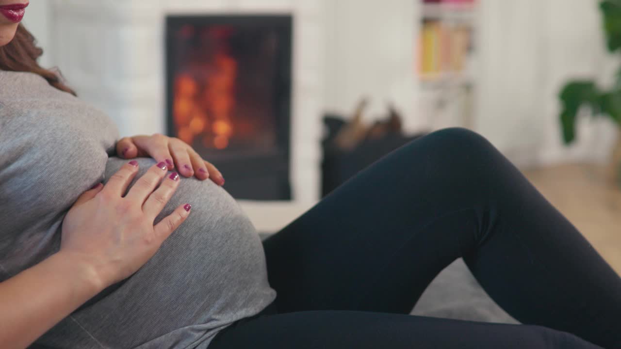 美丽的棕色头发的孕妇坐在舒适的壁炉前抚摸着婴儿的肚子视频素材