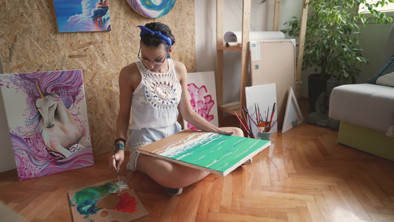 自学成才的年轻女子，一位艺术家，坐在地板上，在画布上画着大海和海滩视频下载