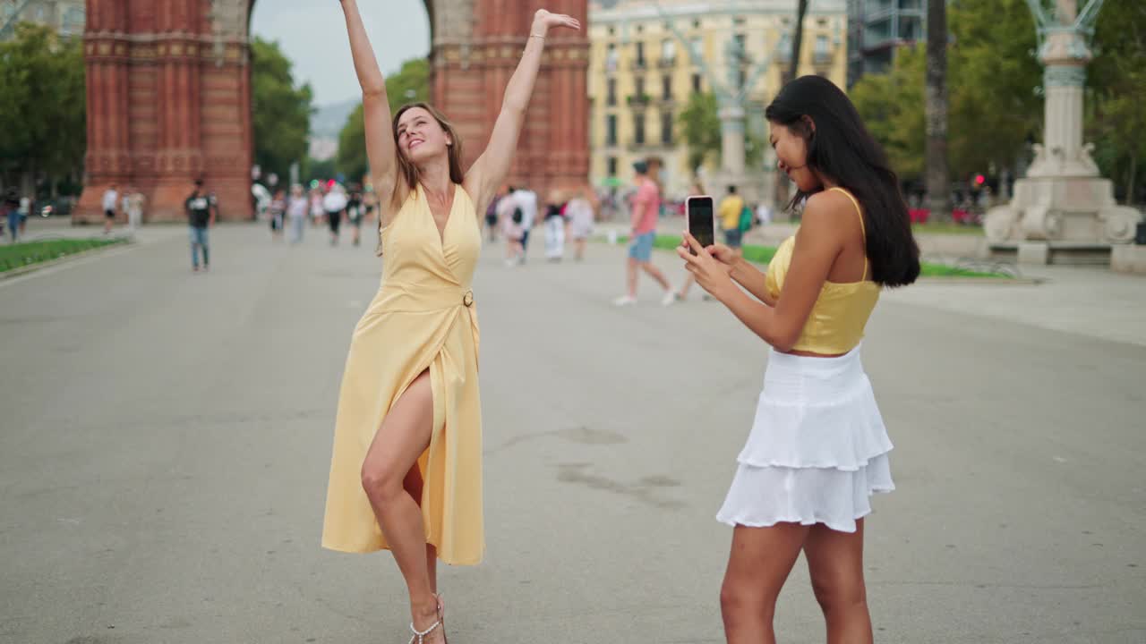年轻的女游客在凯旋门前摆姿势拍照，而她的女性朋友正在拍照视频素材