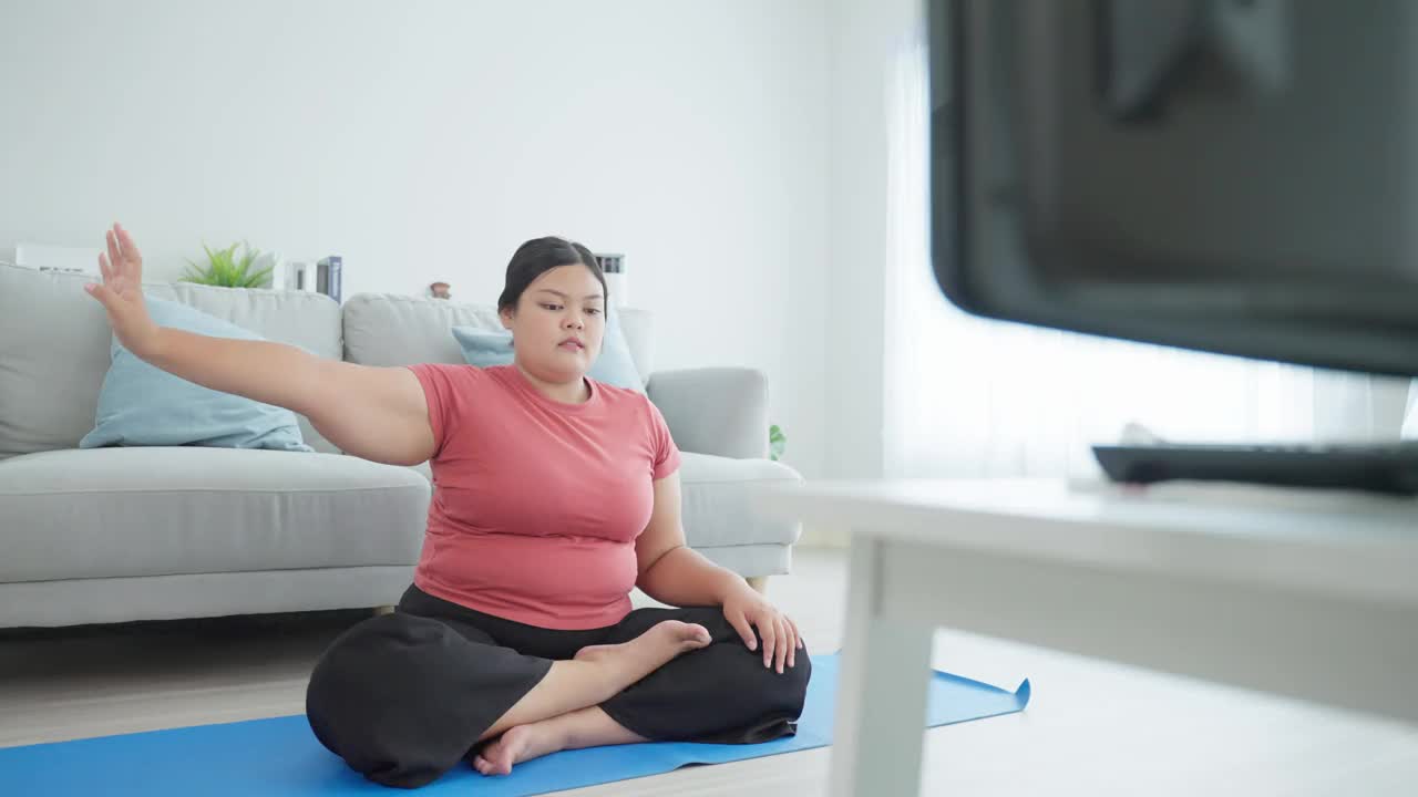 亚洲健美女性在客厅里做瑜伽锻炼。运动女孩坐在自由地板上伸展手，根据在线教练的指导，为健康减肥。视频下载