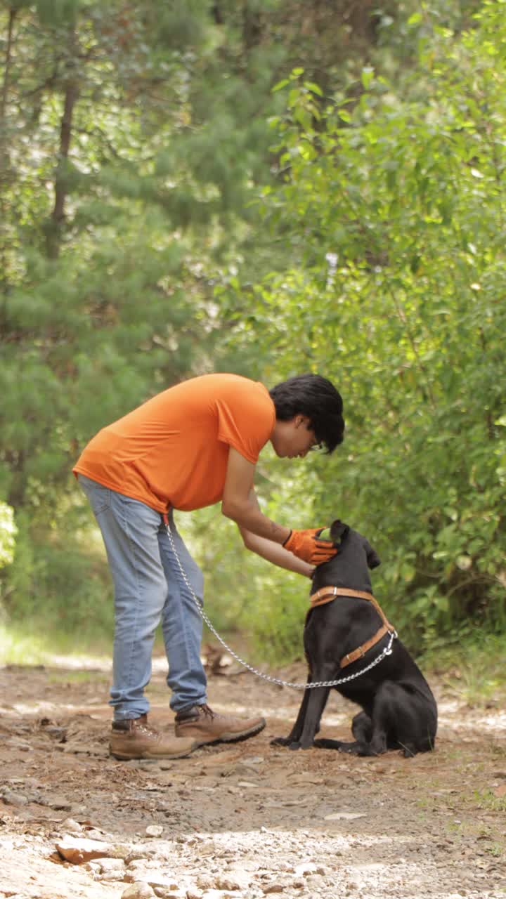 一名身穿橙色t恤的年轻人在森林里抚摸着他的黑狗。视频下载