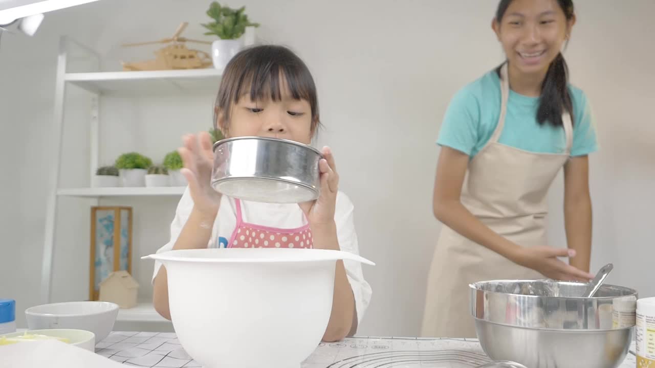 快乐的亚洲孩子在家里一起学习做饼干，女孩教她的姐姐往搅拌碗里撒面粉。视频素材