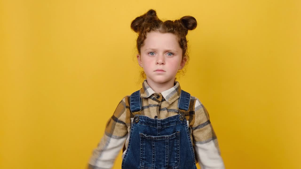 不高兴的、生气的、心烦的小女孩的肖像手拉手交叉放在一边，在画室的黄色背景墙上摆出孤立的姿势。童年情感生活理念视频素材