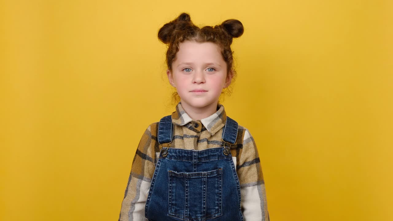 活泼可爱的小女孩的肖像吹送飞吻，穿着衬衫，在画室的黄色墙背景上孤独地摆姿势。童年情感生活理念视频素材
