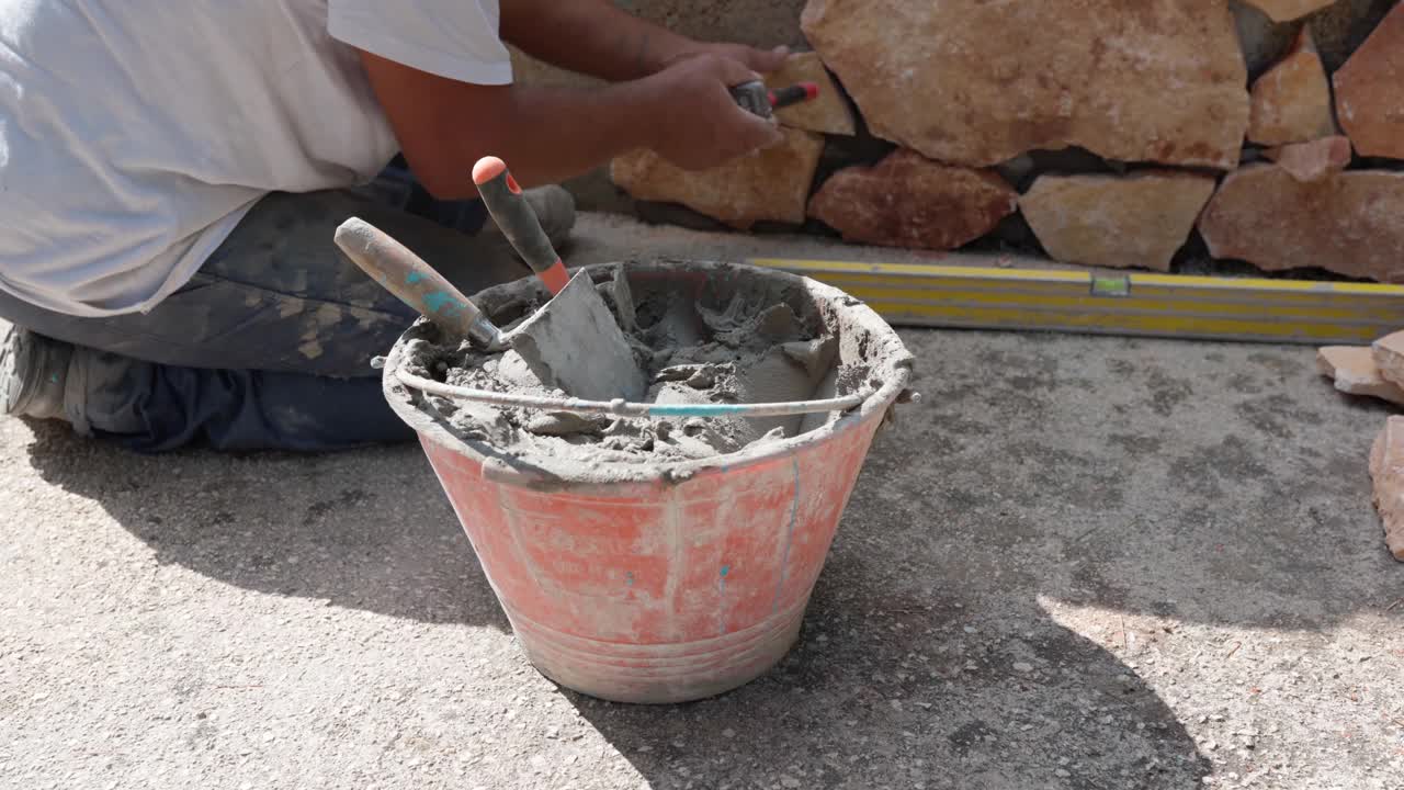 专业工人坐在地上，用水泥溶液将橙色的石头铺在墙面上，砌砖工人用重锤敲打石头并进行找平。修理工工作视频素材