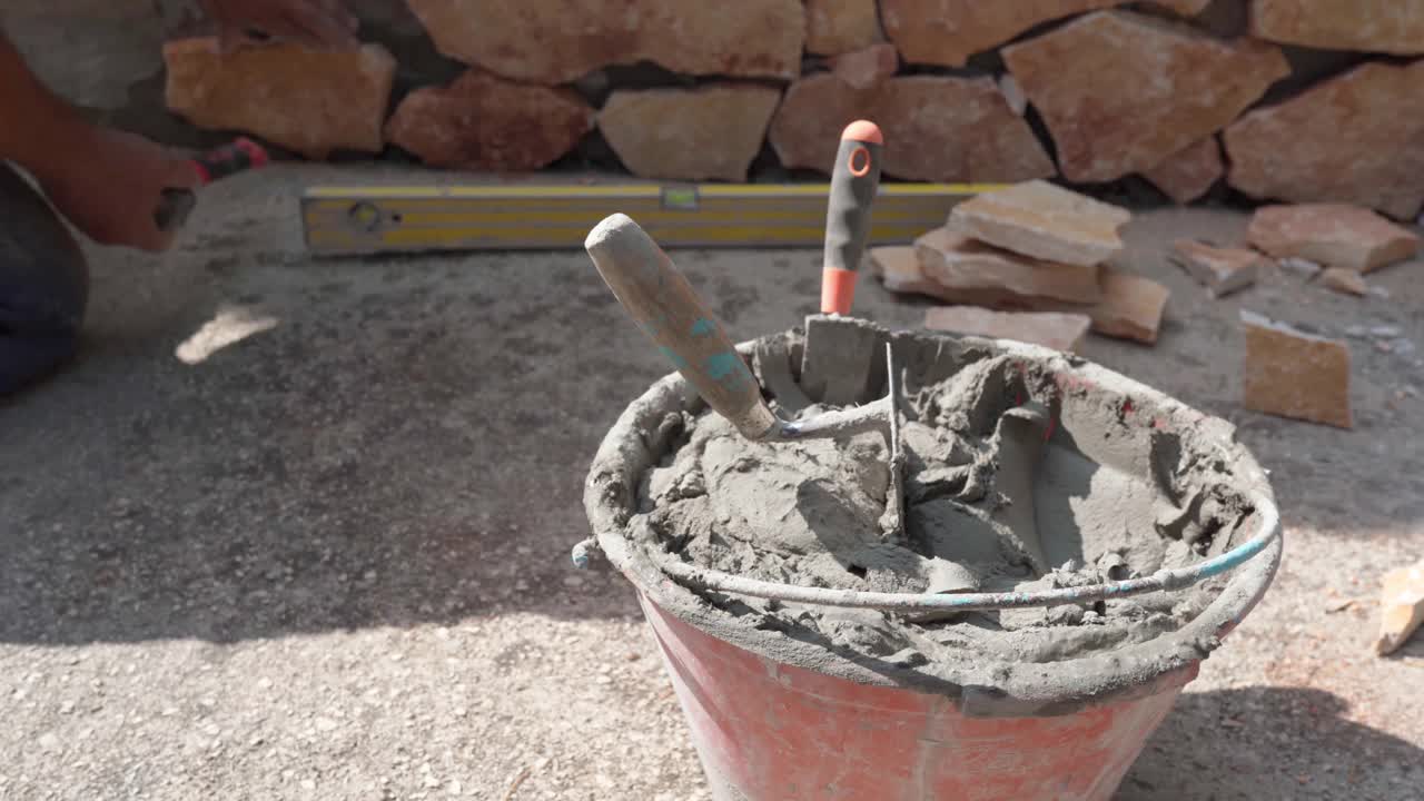 施工现场有装水泥溶液的铲斗、抹刀和镘刀，工人在墙上铺石头，用锤子敲打模糊的背景。施工现场铺设石材的过程视频素材