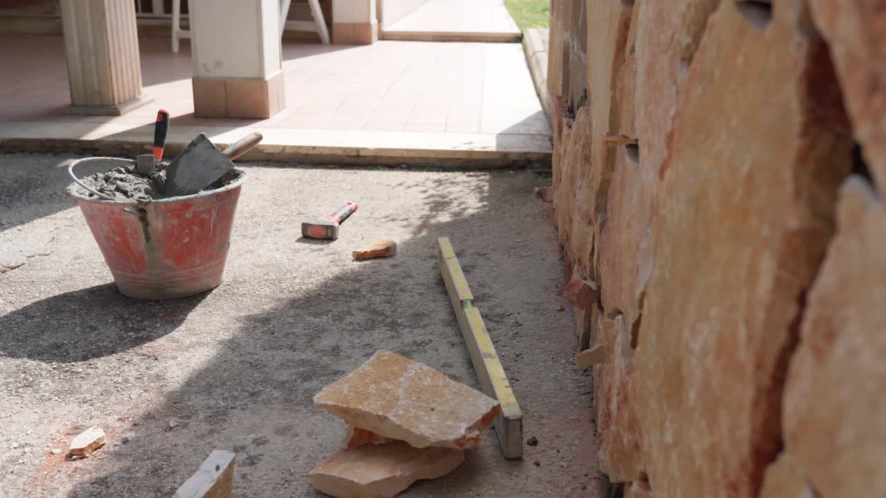 施工现场的施工工具和工具散落在地面上，在装修墙外的水泥溶液上辛苦地铺设橙黄色的石头。外墙维修工程视频素材