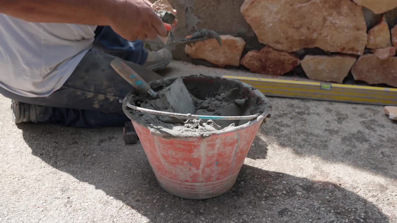 建筑工地上砌砖的工人，不认识的修理工坐在重建墙附近的地上，用金属铲把水泥抹在石头上，铺在墙上视频素材