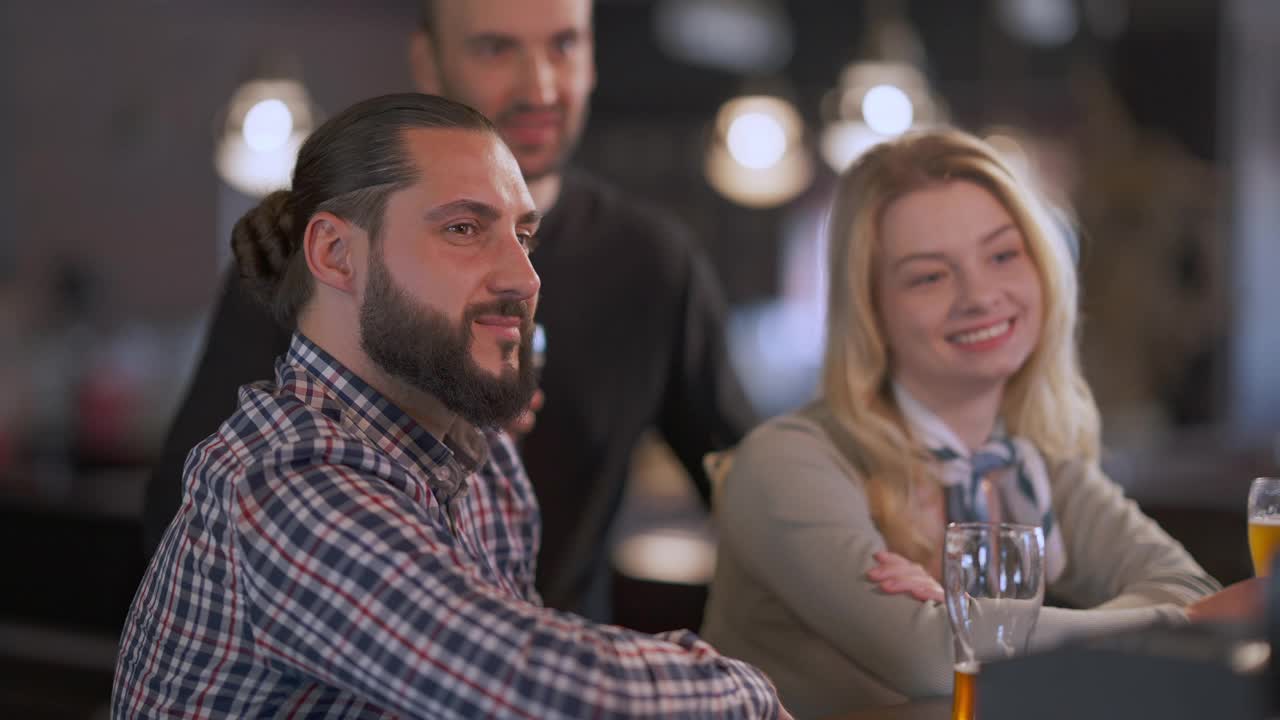 心满意足的一群朋友在酒吧里看电视，坐在吧台边喝着啤酒微笑。积极快乐放松的白种人男人和女人享受休闲周末在酒吧室内饮酒。视频素材