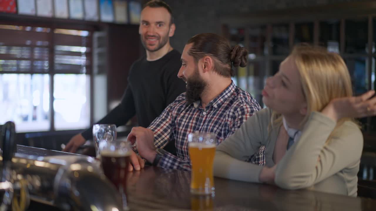 喜气洋洋的白种人夫妇和朋友走进酒吧，坐在吧台边喝啤酒。积极快乐放松的男人和女人在酒吧见面休息的周末。生活方式和友谊。视频素材