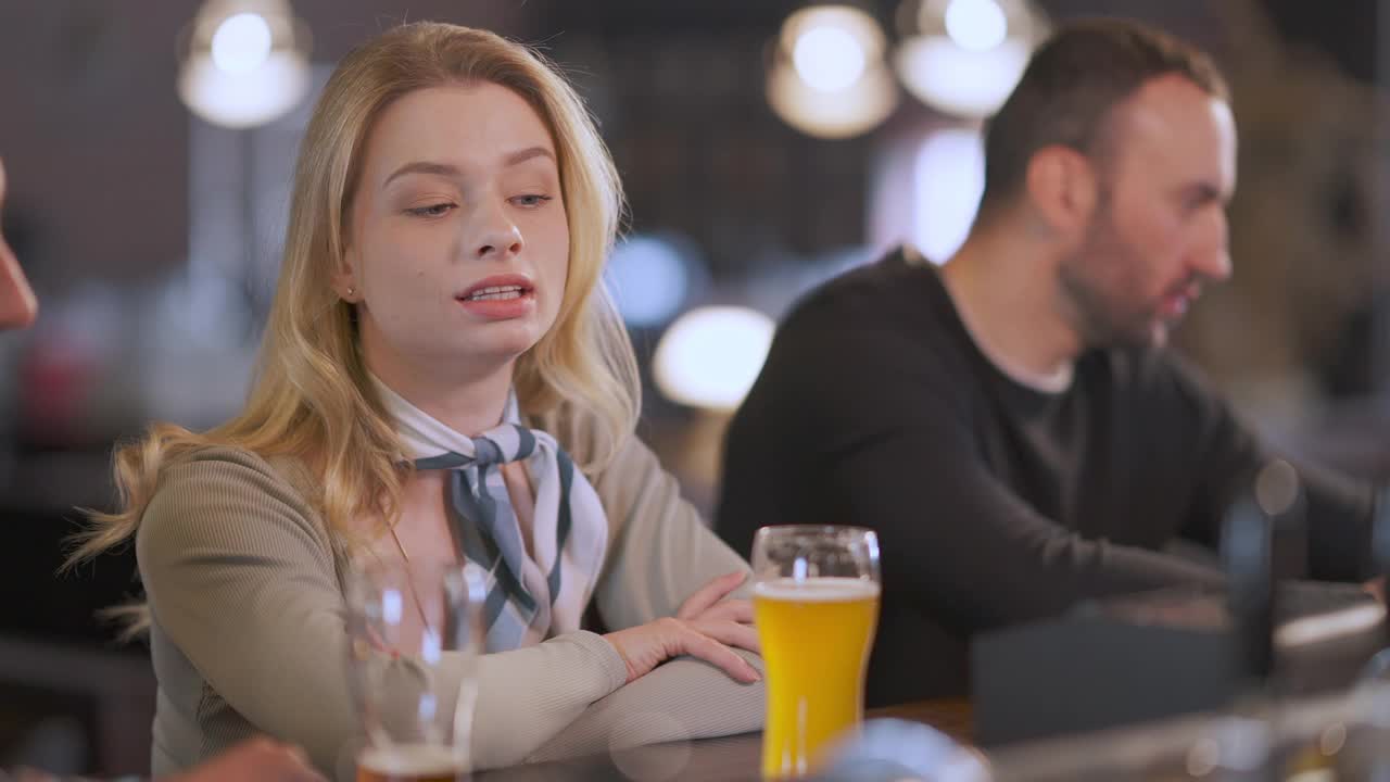 年轻美丽的女人与坐在吧台的男人交谈，背景是喝啤酒的模糊客户。轻松的白人女友和男友在酒吧室内约会。视频素材