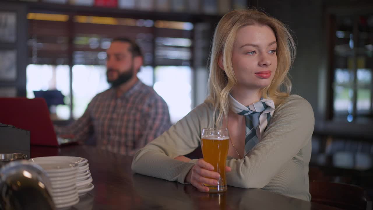 迷人的年轻苗条的女人的肖像坐在吧台与啤酒品脱的男人在后台喝酒。美丽的白种人女士周末在餐厅室内休息。缓慢的运动。视频素材