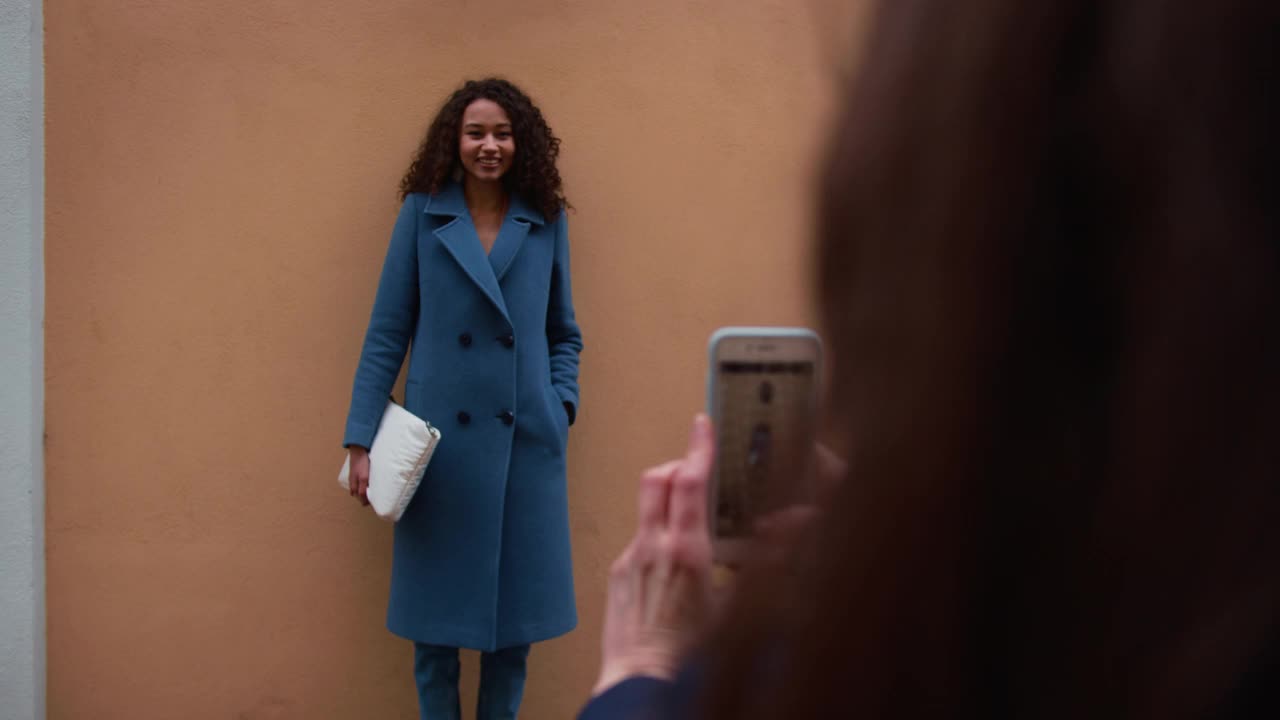 漂亮的年轻快乐的20-25岁的蓝色外套的混血妇女在墙上摆姿势拍照，她的朋友用智能手机视频下载