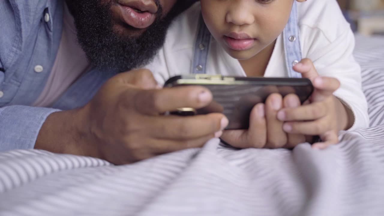 非洲家庭在床上玩手机游戏。父亲和女儿在一起度过了快乐的时光视频素材