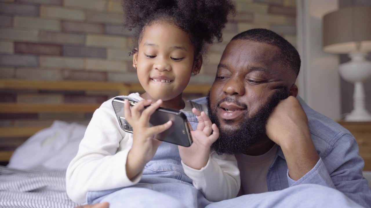 单身黑人父亲和非洲女儿一起躺在床上玩手机app里的视频游戏。周末有活动的家庭视频素材