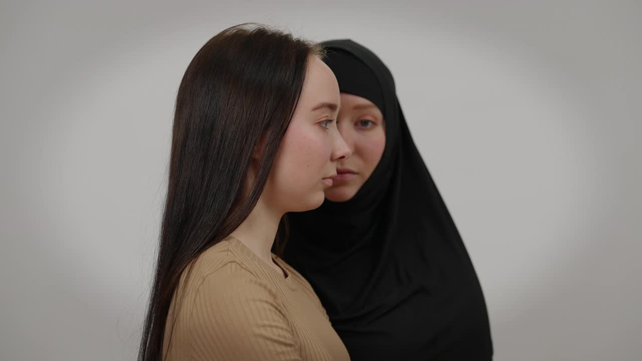 侧视严肃的年轻苗条的女人与模糊的孪生姐妹在黑色头巾的背景。不同宗教信仰的白人女性在灰色背景下摆姿势视频素材