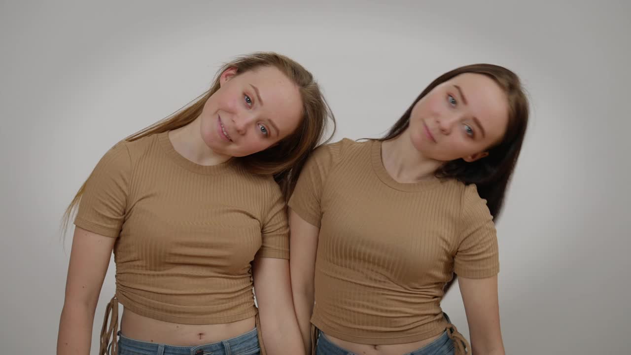 苗条美丽的同卵双胞胎姐妹在灰色背景下同时跳舞看着相机。正面视角中拍的幸福迷人的白人兄弟姐妹微笑视频下载