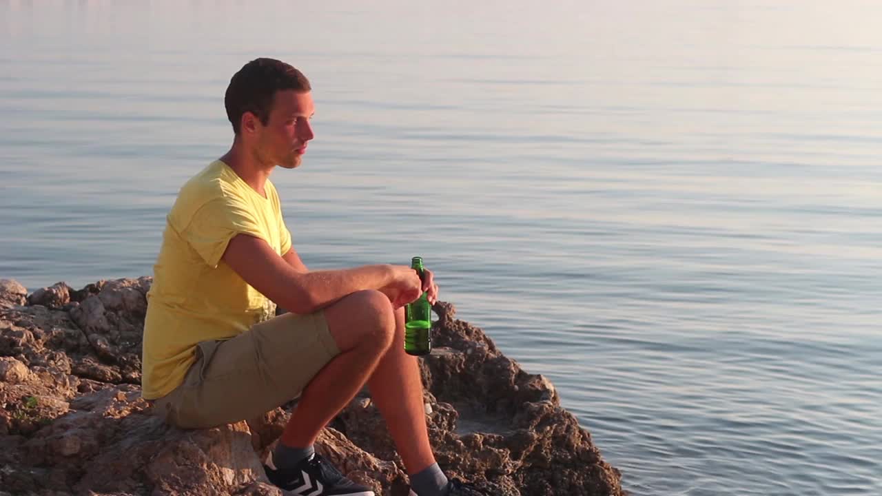 日落时分，一个男人在海边的岩石上喝啤酒视频素材
