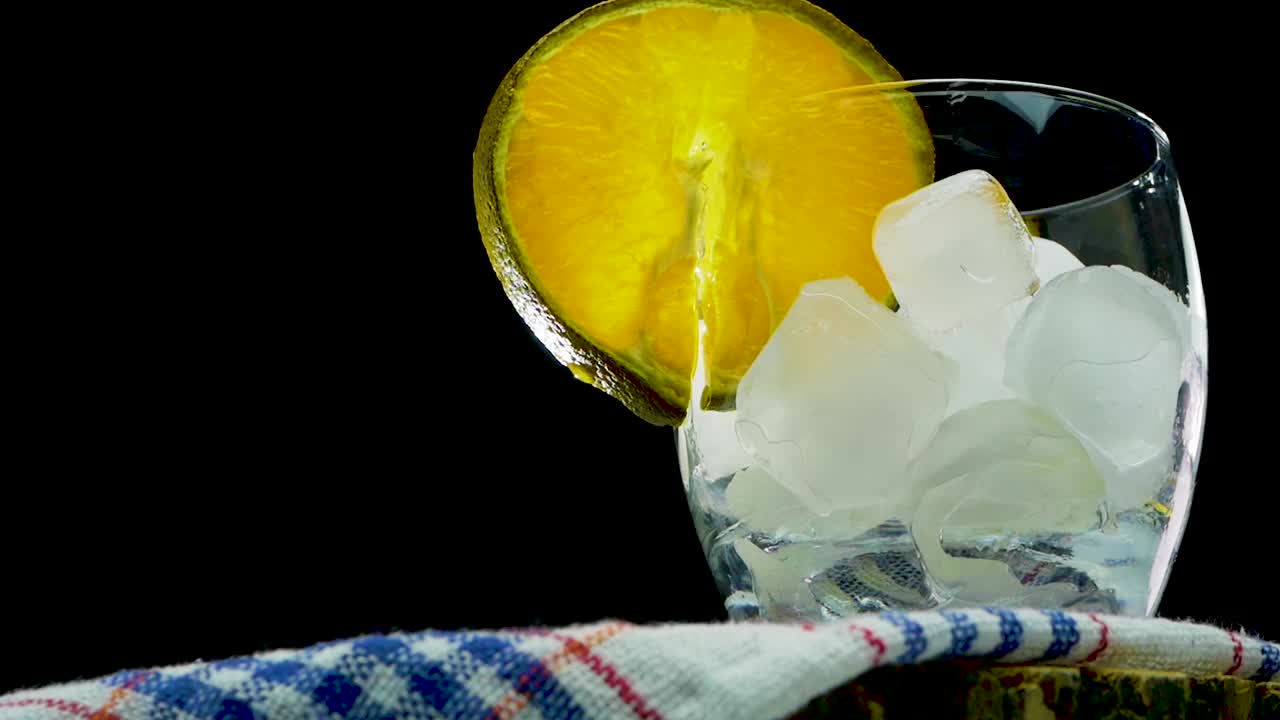 一杯天然橙汁，将橙片放入玻璃杯中，慢动作将黄色柠檬汁倒入黑色背景的玻璃杯中视频素材