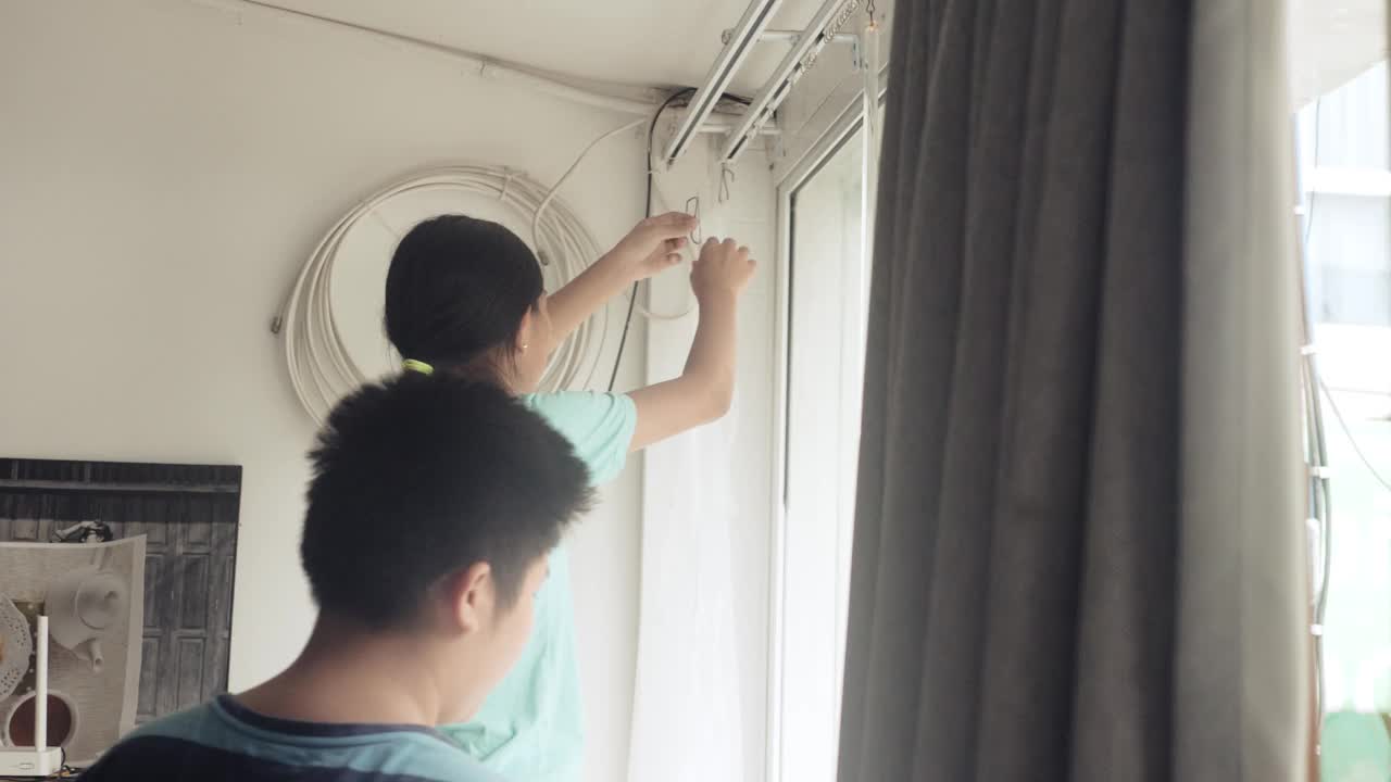 亚洲女孩帮妈妈在家里的窗户旁挂新床单白窗帘，隔离生活理念。视频下载