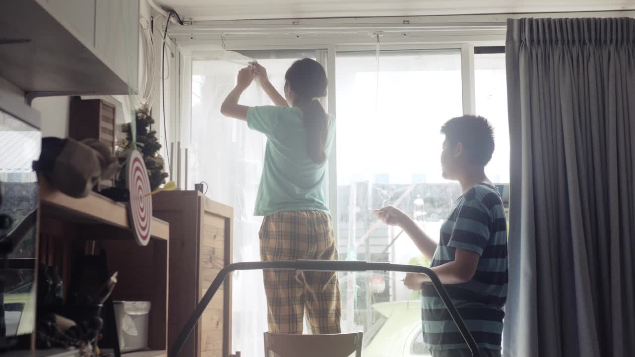亚洲女孩帮妈妈在家里的窗户旁挂新床单白窗帘，隔离生活理念。视频下载