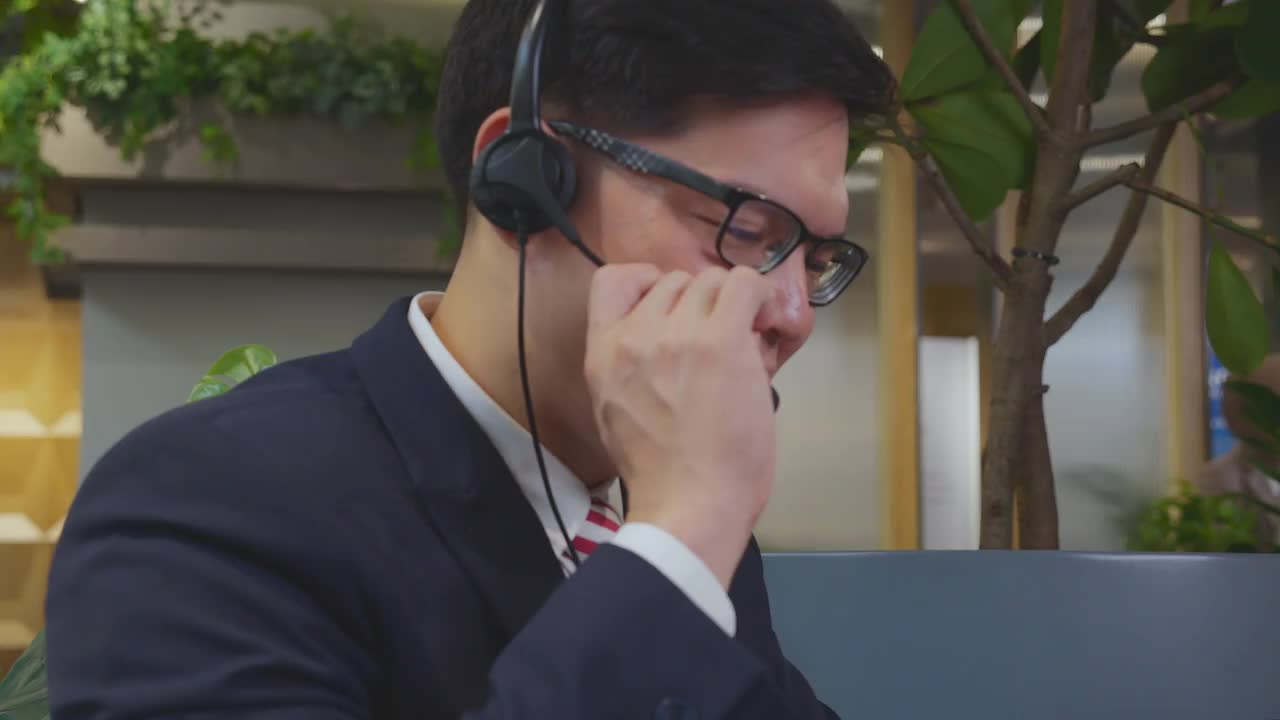 快乐年轻的亚洲商人在耳机和使用笔记本电脑的视频电话会议在现代办公室，男人与耳机交谈，呼叫中心，业务和沟通的概念。视频素材