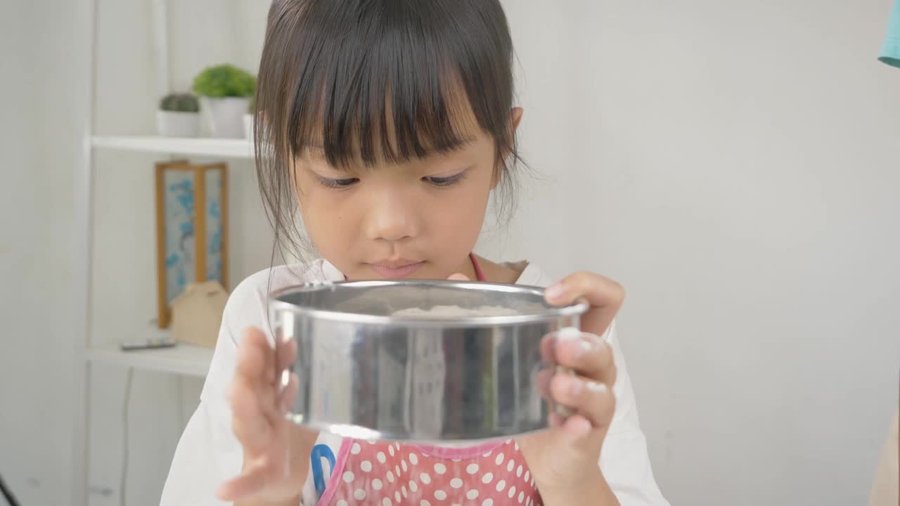 快乐的亚洲孩子在家里学习做饼干，女孩筛面粉到搅拌碗。视频下载