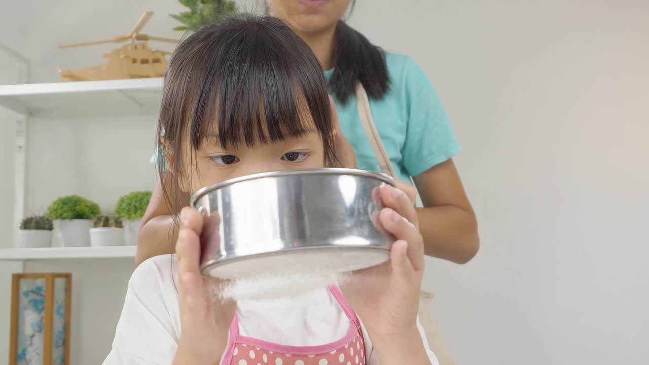 快乐的亚洲孩子在家里学习做饼干，女孩将面粉筛入搅拌碗。视频下载