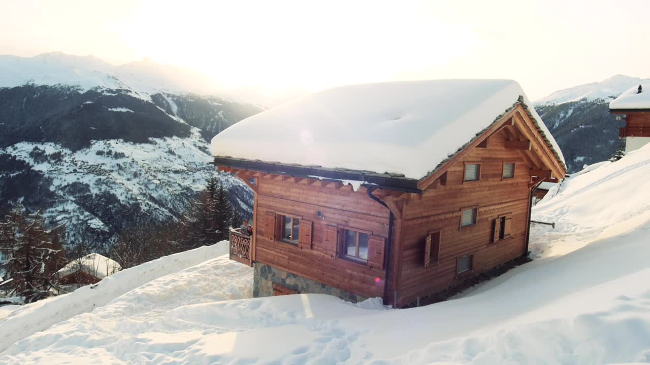 传统的木屋，俯瞰雪山的壮丽景色。阿尔卑斯山冬天的雪。两个孩子在阳台上向镜头挥手。日落的背景。视频素材