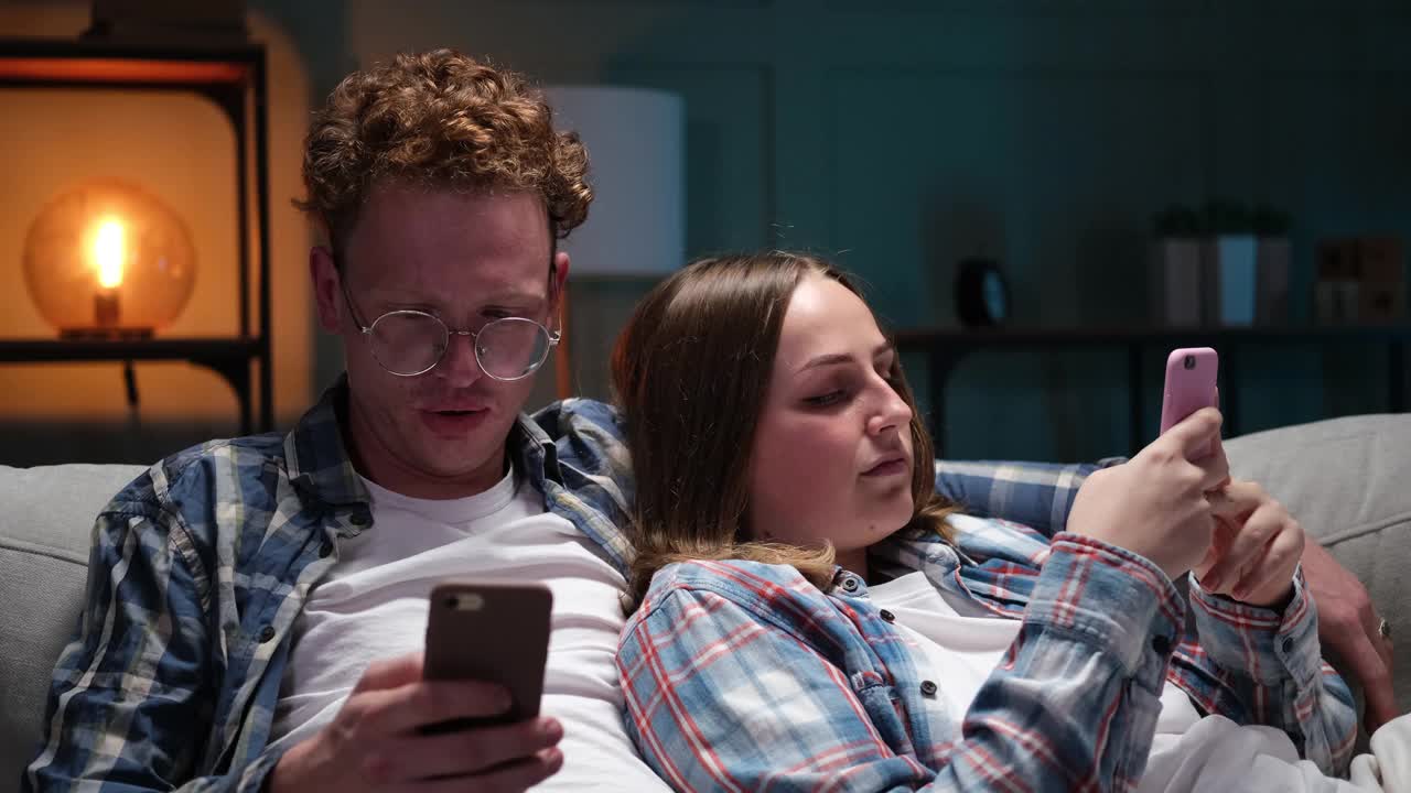 漂亮的年轻夫妇用着智能手机，坐在家里的沙发上视频素材