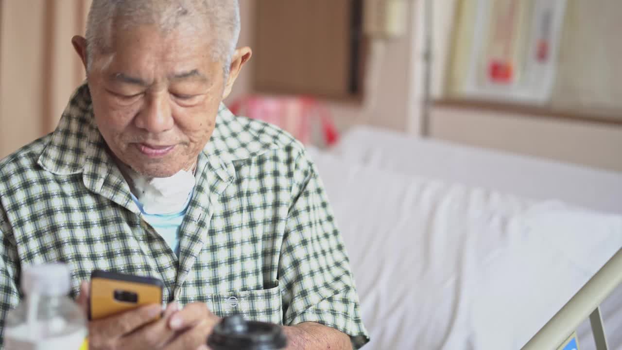 病房里坐在床上看智能手机的亚裔华裔老年男性患者视频下载