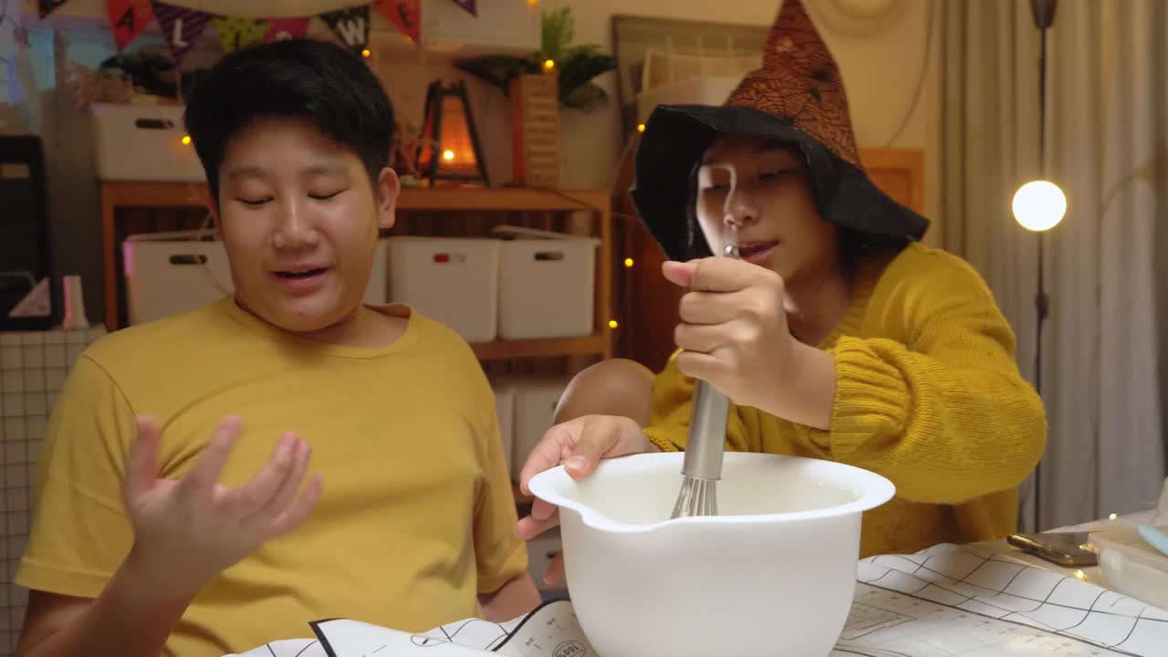 亚洲女孩戴着女巫的帽子，晚上和哥哥一起在家做糖霜饼干，万圣节的生活理念。视频下载