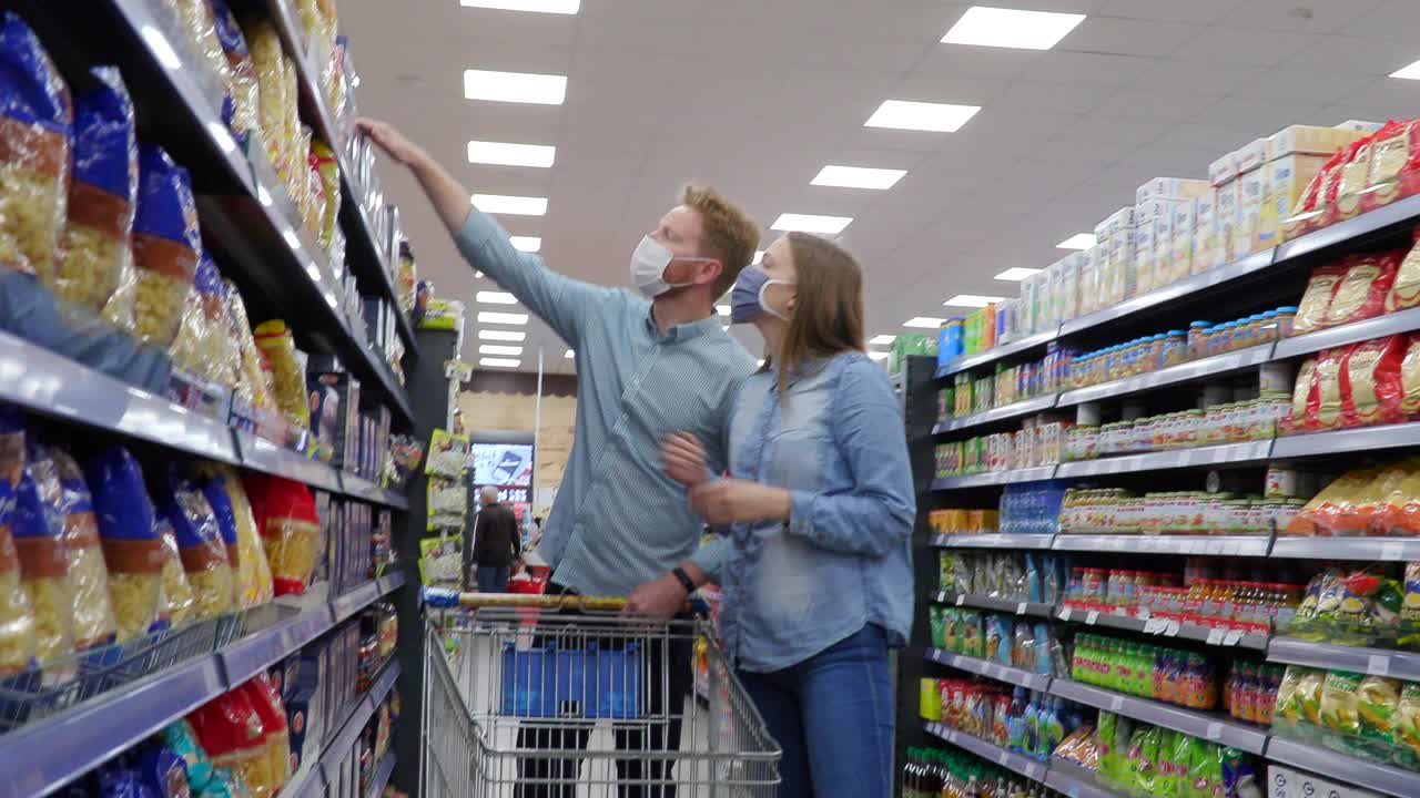 一对戴着口罩的年轻夫妇推着购物车在超市的过道上购买食品视频下载
