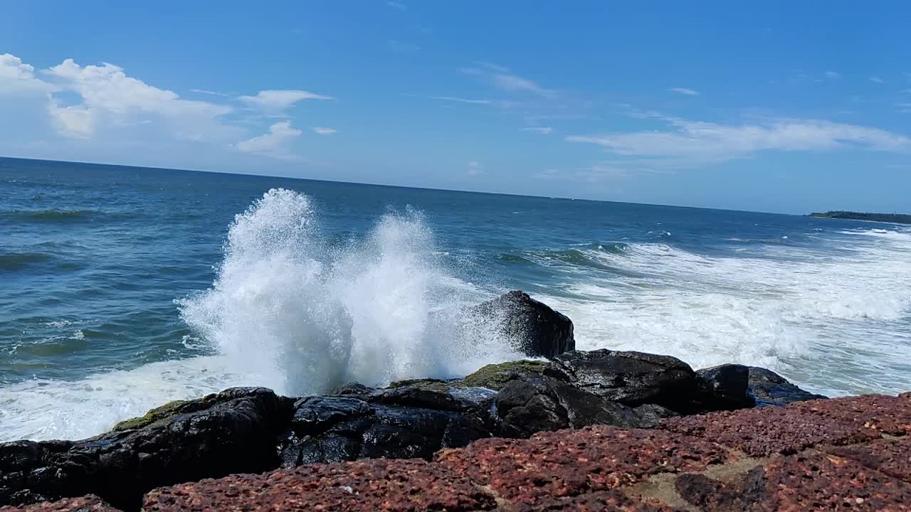 在阳光明媚的日子里，蔚蓝的天空下，白色的海浪拍打着海边的岩石视频下载