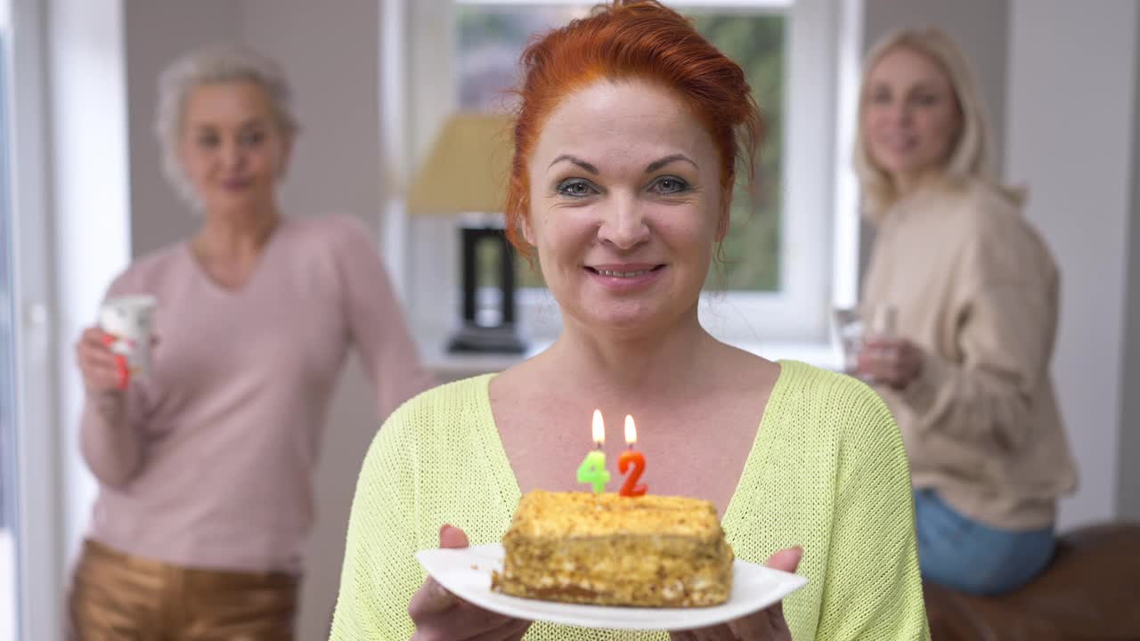 快乐的红发美女拿着生日蛋糕吹灭42年蜡烛，微笑着看着镜头，朋友们在欢呼。欢快自信快乐的女士在家里庆祝视频下载