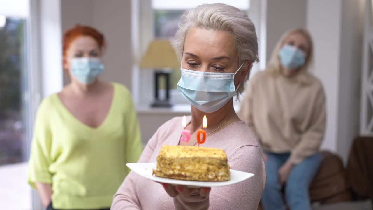一名戴着冠状病毒口罩的白人妇女的肖像，上面挂着蛋糕和50年蜡烛。一名成年女子在Covid-19封锁期间与朋友在家里庆祝节日视频素材