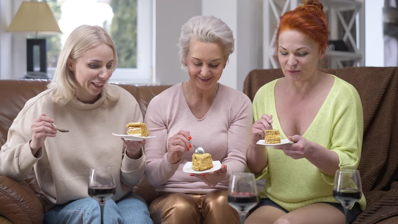 前视图肖像三个快乐的成年白人苗条的妇女品尝美味的蛋糕微笑交谈。放松的朋友坐在客厅的沙发上享受甜点的味道视频素材