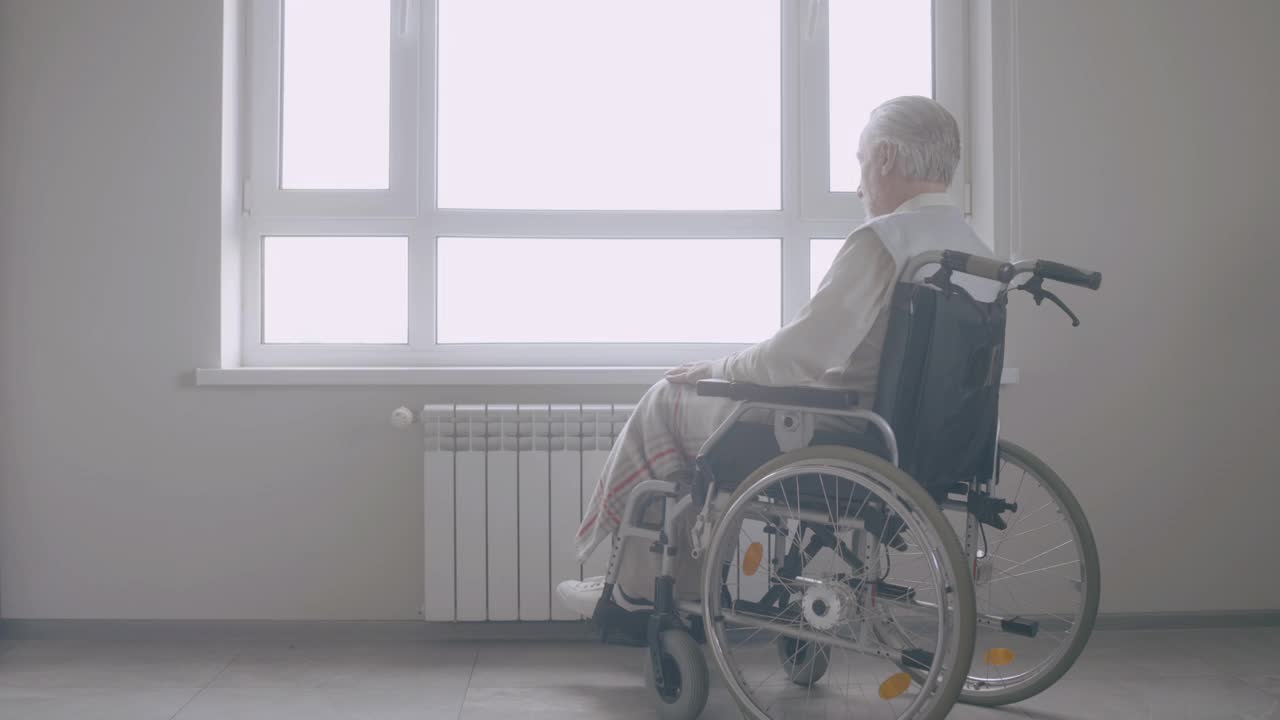 坐在轮椅上的老人悲伤地望着窗外，年老时的孤独视频素材