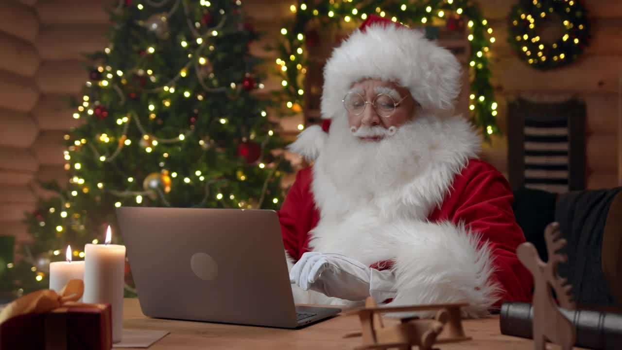 戴着白手套的圣诞老人正坐在他的办公室里，在他的笔记本电脑里键入一些东西视频下载