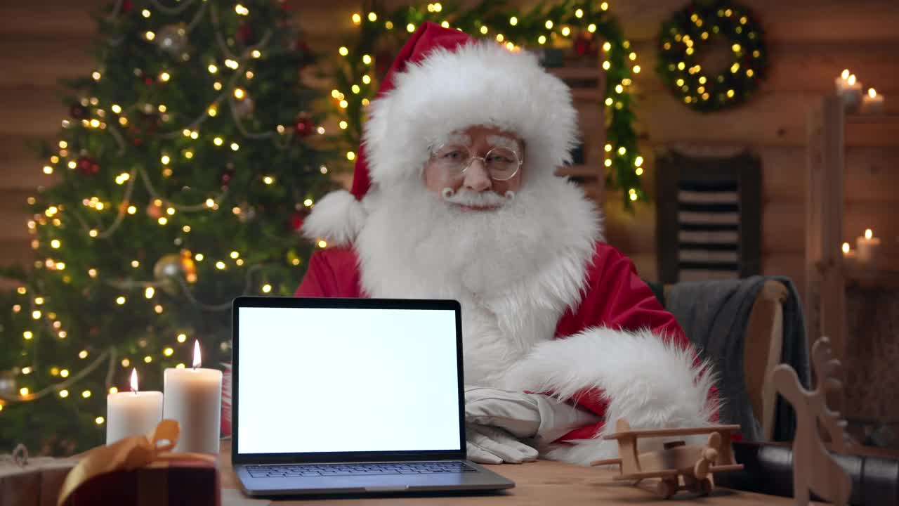 圣诞老人在镜头前竖起大拇指，他面前的桌子上放着一个空的笔记本电脑屏幕视频素材