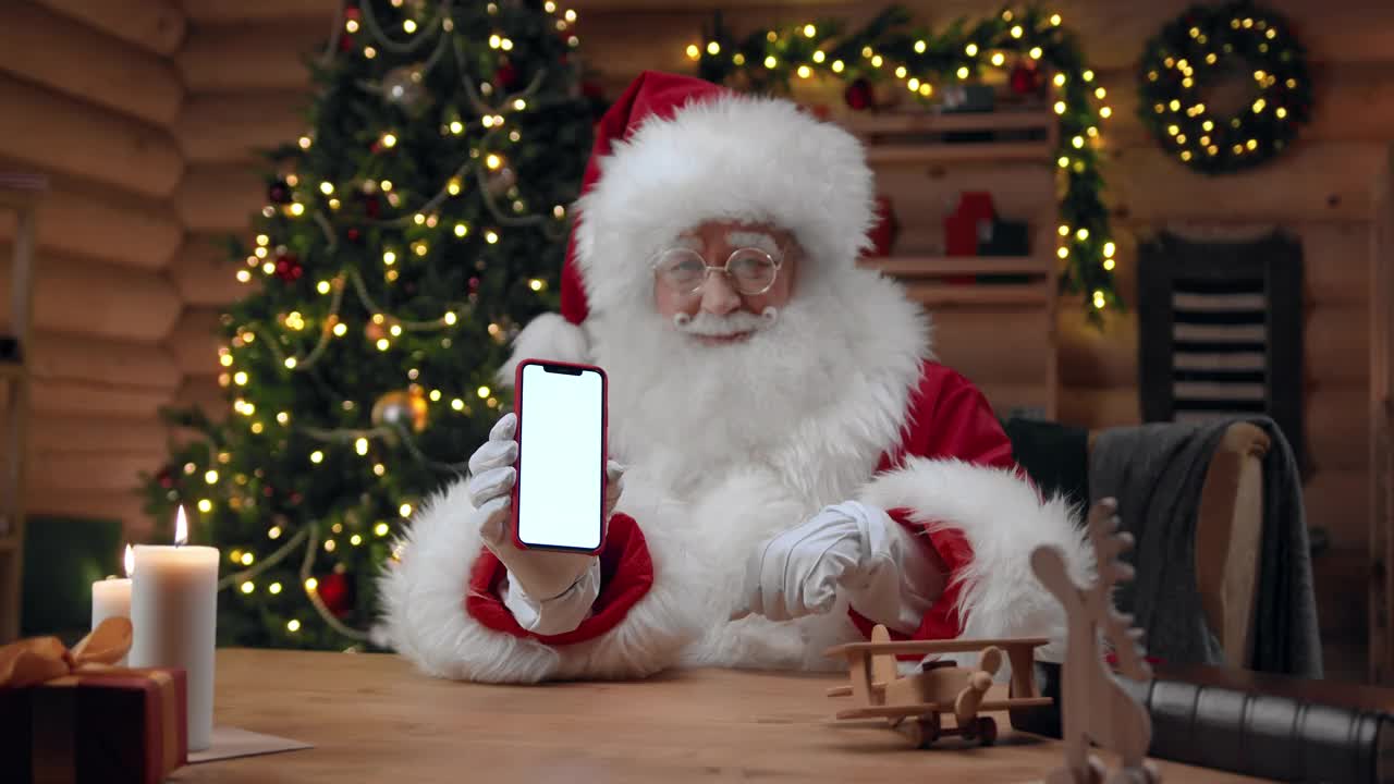 在装饰华丽的室内，圣诞老人展示了一个空屏幕的智能手机，并微笑着向相机挥手视频下载