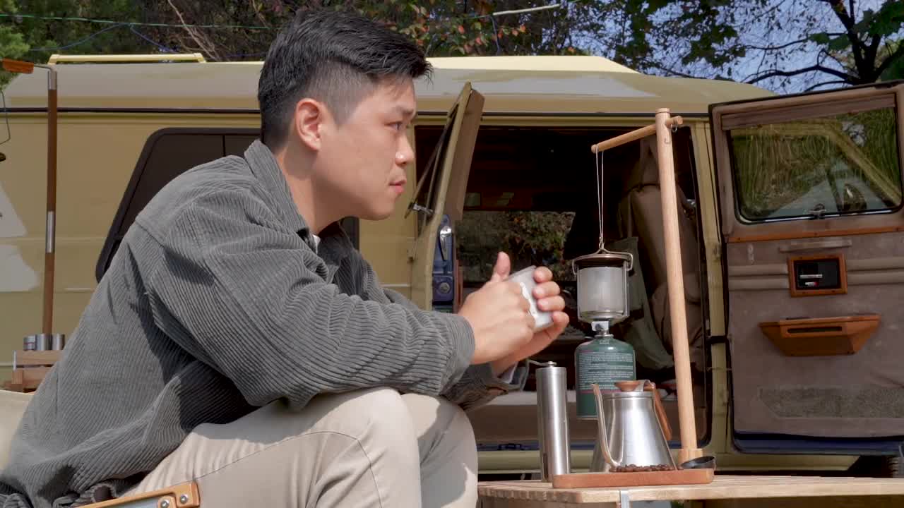 户外露营-年轻人坐在帐篷前的桌子旁享受放松，一边喝着不锈钢杯的咖啡视频下载