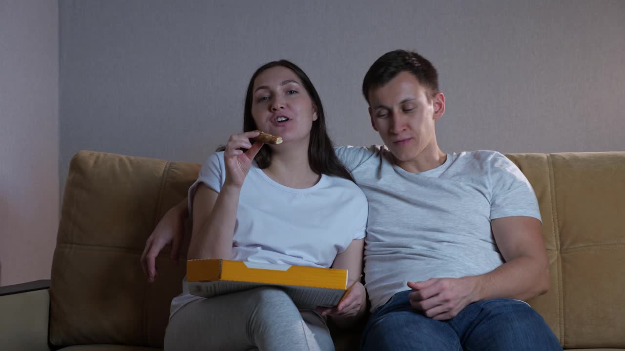 男人和女人坐在沙发上吃比萨饼和看电视视频素材