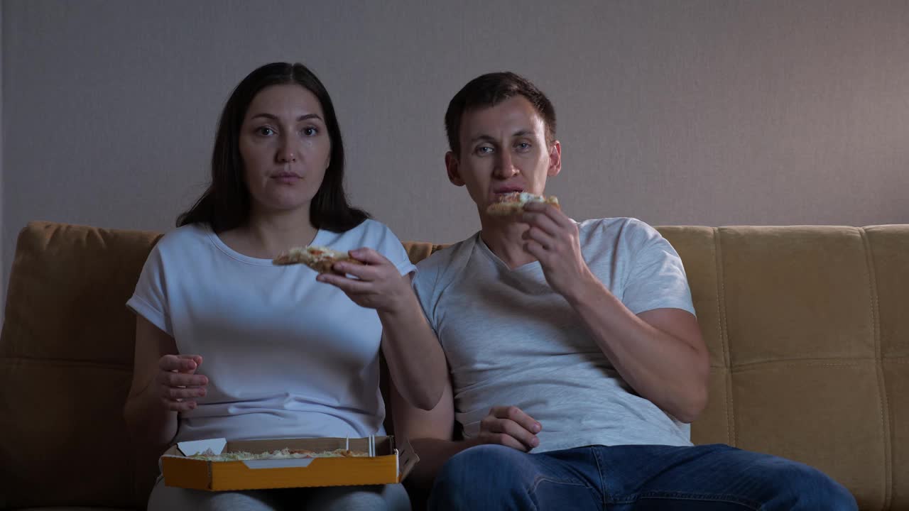 男人和女人在猫旁边的沙发上吃披萨看电视视频素材